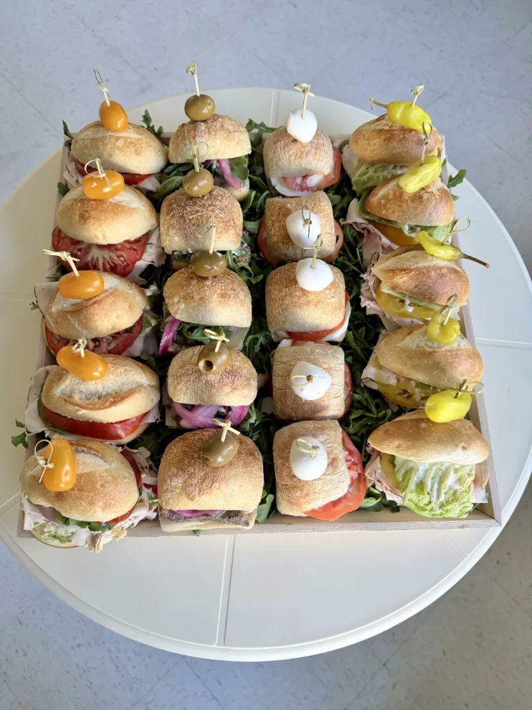 Top-down view of a luxury corporate catering tray filled with artisanal mini sliders on craft rolls, topped with olives, cherry tomatoes, mozzarella, and pepperoncini over fresh greens.