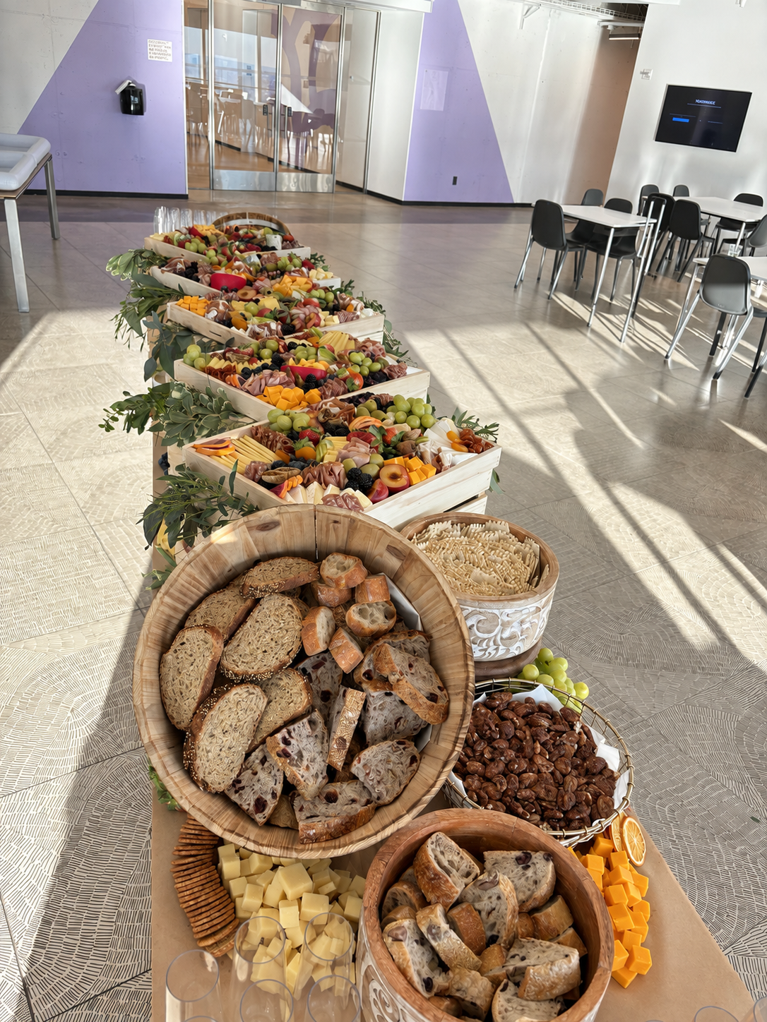 An opulent long corporate catering buffet featuring wooden bowls of artisanal sliced sourdough breads, roasted nuts, cubed cheeses, and tiered wooden crates filled with lavish charcuterie, fresh fruits, and cured meats in a modern executive event venue.