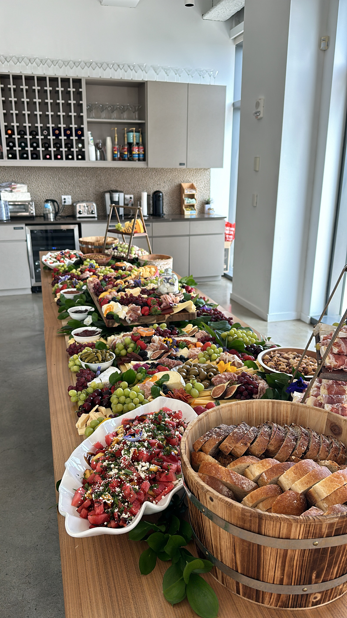 An expansive luxury corporate grazing table spread on a long wooden table in a modern corporate pantry, featuring a wooden bucket filled with sliced artisan bread, a fresh watermelon feta salad, assorted charcuterie, fine cheeses, fresh fruits, olives, and berries.