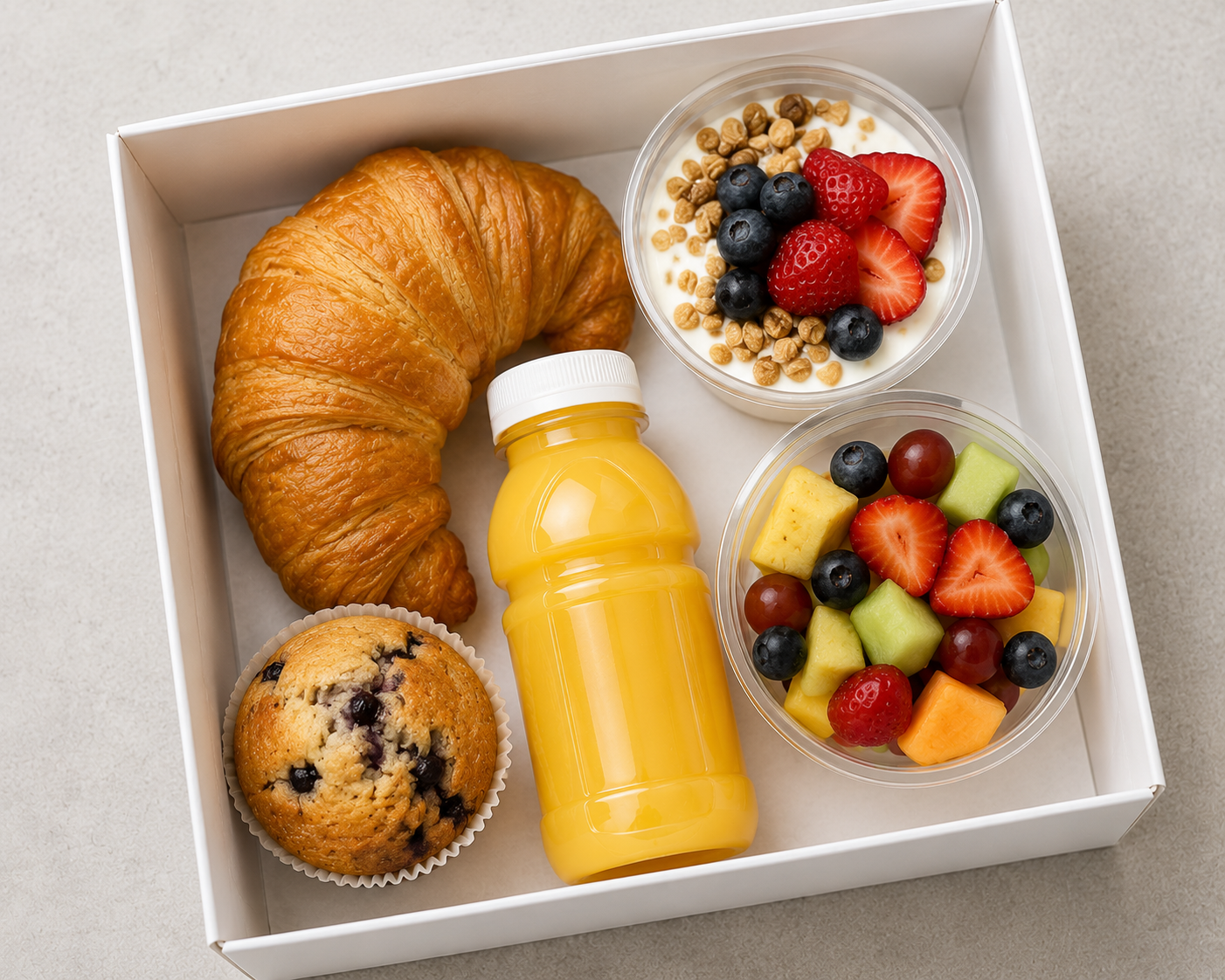 An executive breakfast catering box featuring a golden butter croissant, blueberry muffin, fresh-squeezed orange juice, yogurt parfait with berries, and fresh fruit salad.
